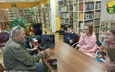 W bibliotece przy stole siedzi młodzież z Piotrem Martinem. Piotr pokazuje im kamerę, ma otwarte laptopa i pokazuje im jak nagrywa się kr&oacute;tkie filmiki.