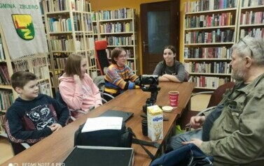 Przy stole w bibliotece siedzi młodzież i Piotr Martin, na stole stoi kamera na stojaku, akcesoria do tego i karton mleka.