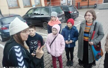 Na parkingu przy Urzędzie Gminy stoi Iwona Skwarek z młodzieżą im coś pokazując.