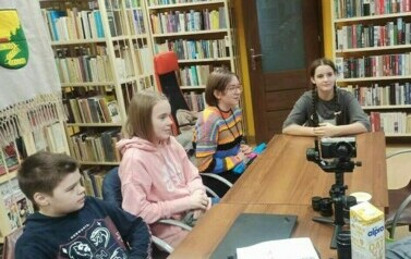 Młodzież siedzi przy stole w bibliotece, na stole stoi kamera na stojaku i akcesoria do tego.