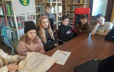 W bibliotece przy stole siedzi grupa mlodzieży i słucha Piotra Martina. Na stole leży kartka i długopis i  mała torba.