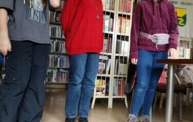 W bibliotece stoją trzy dziewczyny, a przed nimi na podłodze leżą trzy maskotki.