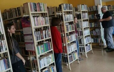 Piotr jest schowany w bibliotece z kamerą pomiędzy p&oacute;łkami i nagrywa trzy dziewczyny, kt&oacute;re też się za regałami chowią pokazując tylko kawałek swojej twarzy.