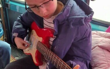 Chłopiec siedzi w busie trzymając gitarę elektryczną i pr&oacute;bując grać.