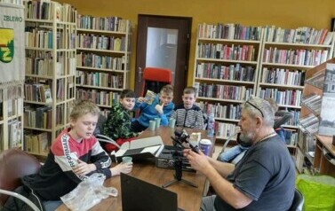 Przy stole w bibliotece siedzi Piotr Martin  z chłopccami i pokazuje im działanie kamery.