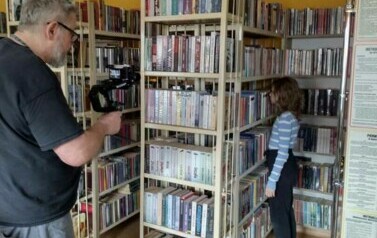 Pomiędzy regałami w bibliotece stoi dziewczynka, a w pobliżu stoi Piotr Martin i ją nagrywa kamerką.