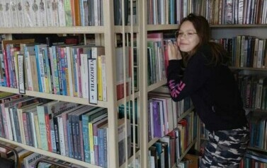 Pomiędzy regałami w bibliotece jest schowana dziewczynka w okularach.