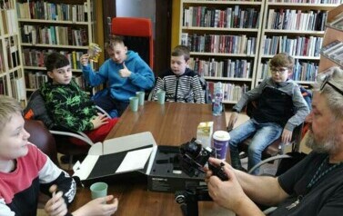 Przy stole w bibliotece siedzi młodzież i Piotr Martin, na stole stoi kamera na stojaku, akcesoria do tego.