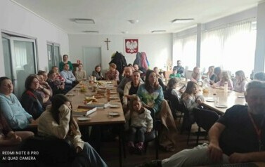 Przy stołach na białej sali siedzą licznie rodzice i ich dzieci oglądając nagrania filmowe i muzyczne dzieci.