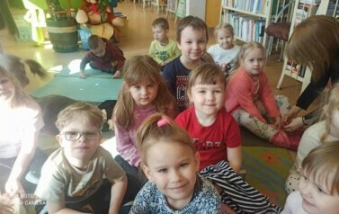 Grupa dzieci siedzących na dywanie. Widać nauczycielkę, kt&oacute;ra poprawia coś przy bucie dziewczynki.