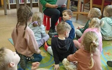 Dzieci siedzą na dywanie i patrzą w stronę bibliotekarki, kt&oacute;ra czyta im zagadki.