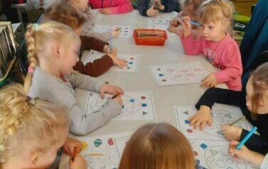 Dzieci przy stole uzupełniają kredkami świąteczne karty pracy. 