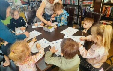 Dzieci przy stole uzupełniają kredkami świąteczne karty pracy. Nad stołem pochylone są ich Panie pomagając im.