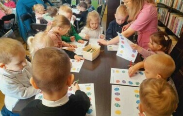 Dzieci przy stołach uzupełniają kredkami świąteczne karty pracy. Nad stołami pochylone są ich Panie pomagając im.