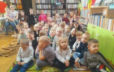 Na dywanie siedzą dzieci i słuchają bajki czytanej przez bibliotekarkę.Z tyłu na krzesełku siedzi ich Pani wraz z dziewczynką na kolanach.