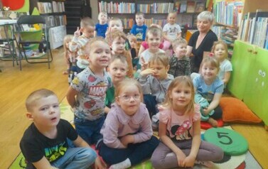 Dzieci siedzą na dywanie wraz ze swoją Panią i słuchają bajki czytanej przez bibliotekarkę.