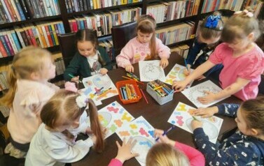 Dziewczynki siedzą przy stole i uzupełniają kredkami świąteczne karty pracy.