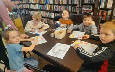 Dzieci siedzą przy stole i uzupełniają kredkami świąteczne karty pracy. Przy stole widać Panią opiekunkę.