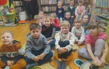 Dzieci siedzą na dywanie i słuchają bajki, kt&oacute;rą czyta im bibliotekarka.