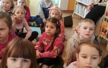 Dzieci siedzą na dywanie i słuchają bajki, kt&oacute;rą czyta im bibliotekarka.