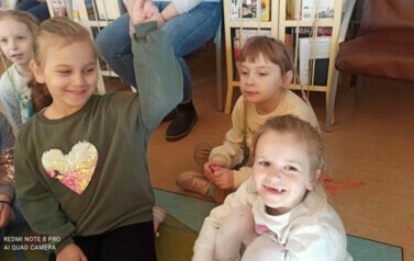 Uśmiechnięte dzieci siedzą na dywanie i słuchają bajki, kt&oacute;rą czyta im bibliotekarka.