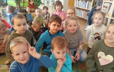 Dzieci siedzą na dywanie i słuchają bajki, kt&oacute;rą czyta im bibliotekarka.