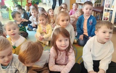 Dzieci siedzą na dywanie i słuchają bajki, kt&oacute;rą czyta im bibliotekarka.