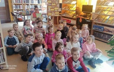Dzieci siedzą  na dywanie. Za nimi po środku siedzi bibliotekarka z książeczką w dłoniach pokazując ją.