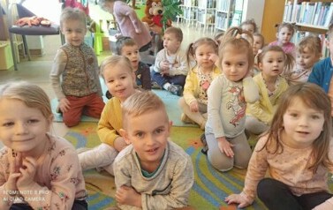 Dzieci siedzą na dywanie i słuchają bajki, kt&oacute;rą czyta im bibliotekarka.