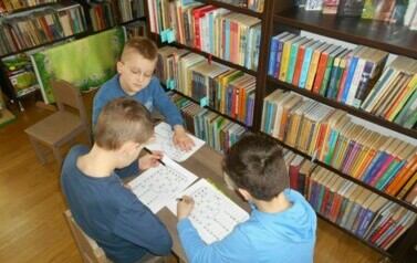 Trzech chłopc&oacute;w przy małym stoliku uzupełniają karty pracy.