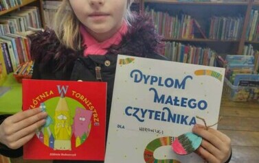 Dziewczynka w jasnej czapce stoi z dyplomem w ręku i wypożyczoną książeczeką.