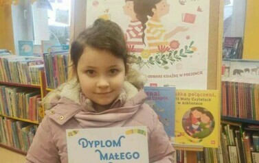 Dziewczynka w ciemnych włosach trzyma dyplom i kredki. Za nią jest tablica korkowa z plakatem promującym projekt Mała Książka Wielki Człowiek.