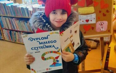 Dziewczynka w czerwonej czapce trzyma dyplom, kredki i tekturową kartkę z grą. Za nią tablica korkowa z walentynową grafiką.