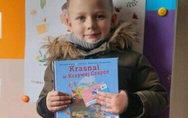 Chłopiec w blond wlosach trzyma książkę z wyprawki. Za nim jest tablica metalowa z plakatem dotyczącym projektu czytelniczego.