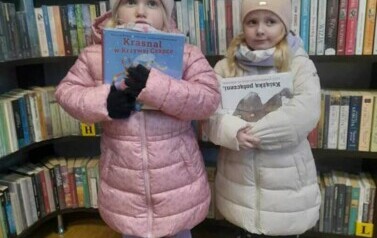 Dwie dziewczynki stoją z  książeczkami w rękach pokazując je. Za nimi są regały z książkami.