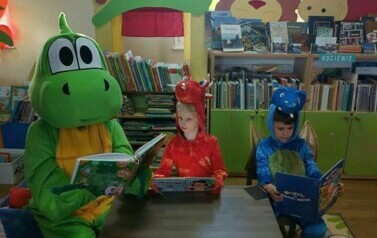 Duży smok z małymi dwoma smokami siedzą przy małym stoliku i czytają bajki.