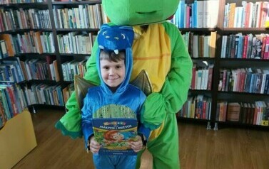 Duży zielony smok stoi z małym niebieskim smokiem, kt&oacute;ry trzyma książeczkę o zwierzątkach.