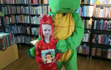 Duży zielony smok stoi z małym czerwonym smokiem, kt&oacute;ry trzyma książeczkę o jeżyku.