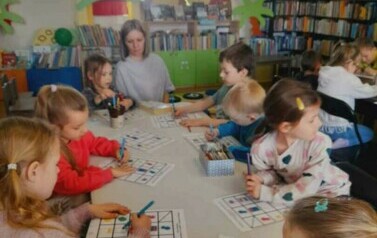 Dzieci siedzą przy stole i robią karty pracy o książce.Na końcu stołu siedzi Pani z Przedszkola.