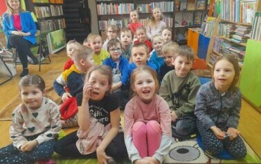 Dzieci siedzą na dywanie i słuchają bajki czytanej przez bibliotekarkę.