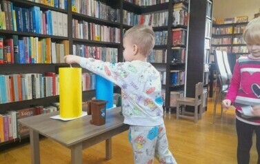 Chłopiec stoi przy stoliku na kt&oacute;rym są zabawkowe kosze na śmieci i wkłada karteczkę z rysunkiem śmiecia do ż&oacute;łtego kosza.