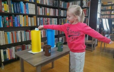 Dziewczynka w r&oacute;żowej bluzce stoi przy stoliku na kt&oacute;rym są zabawkowe kosze na śmieci i wkłada karteczkę z rysunkiem śmiecia do ż&oacute;łtego kosza.