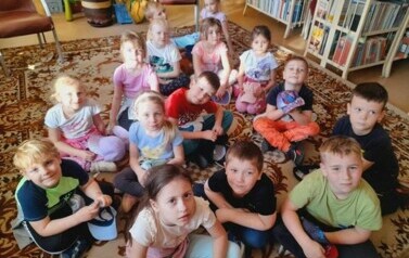 Dzieci siedzą na dywanie i słuchają bajki, kt&oacute;rą czyta im bibliotekarka.