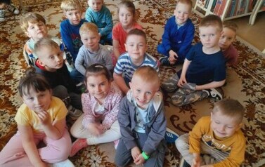 Dzieci siedzą na dywanie i słuchają bajki, kt&oacute;rą czyta im bibliotekarka.