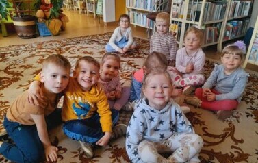Dzieci siedzą na dywanie i słuchają bajki, kt&oacute;rą czyta im bibliotekarka.