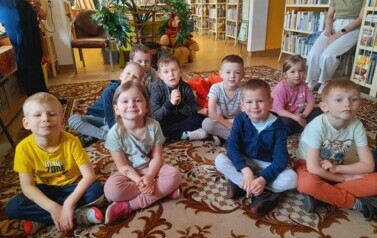 Dzieci siedzą na dywanie i słuchają bajki czytanej przez bibliotekarkę. 
