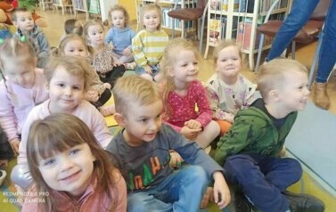 Grupa dzieci siedzi na dywanie i słucha bajki, kt&oacute;rą czyta bibliotekarka.