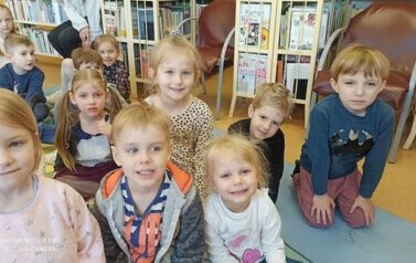 Grupa dzieci siedzi na dywanie i karimatach i słucha bajki czytanej przez bibliotekarkę.