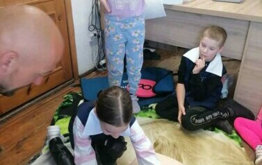 Dziewczynka pochylona jest nad manekinem dotykając jego bark&oacute;w, obok stoi inna dziewczynka. Z prawej strony siedzi chłopiec i patrzy, a z lewej strony siedzi strażak i obserwuje.