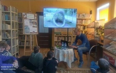 Młodzież siedzi a dywanie, przy małym okrągłym stolku siedzi autor i wyświetla zdjęcia na ekranie telewizora.